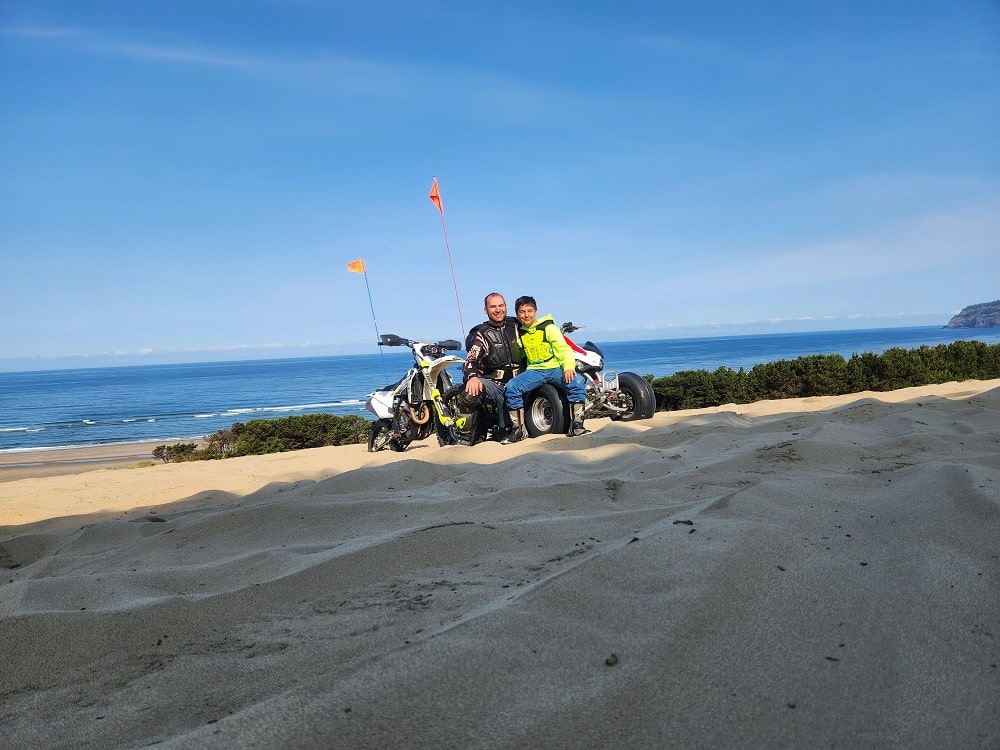 Joe Gambino and his son sitting together on the Oregon Dunes with a Husqvarna FC450 dirt bike and the Pacific Ocean in the background