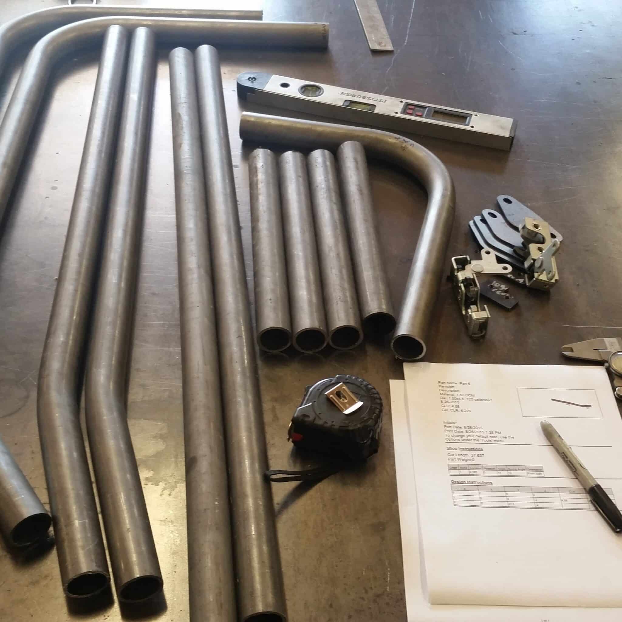DOM steel cage tubes, pre-bent hoop, floor plates, tape measure, digital level, and Bend-Tech part manufacturing instructions laid out before a cage build